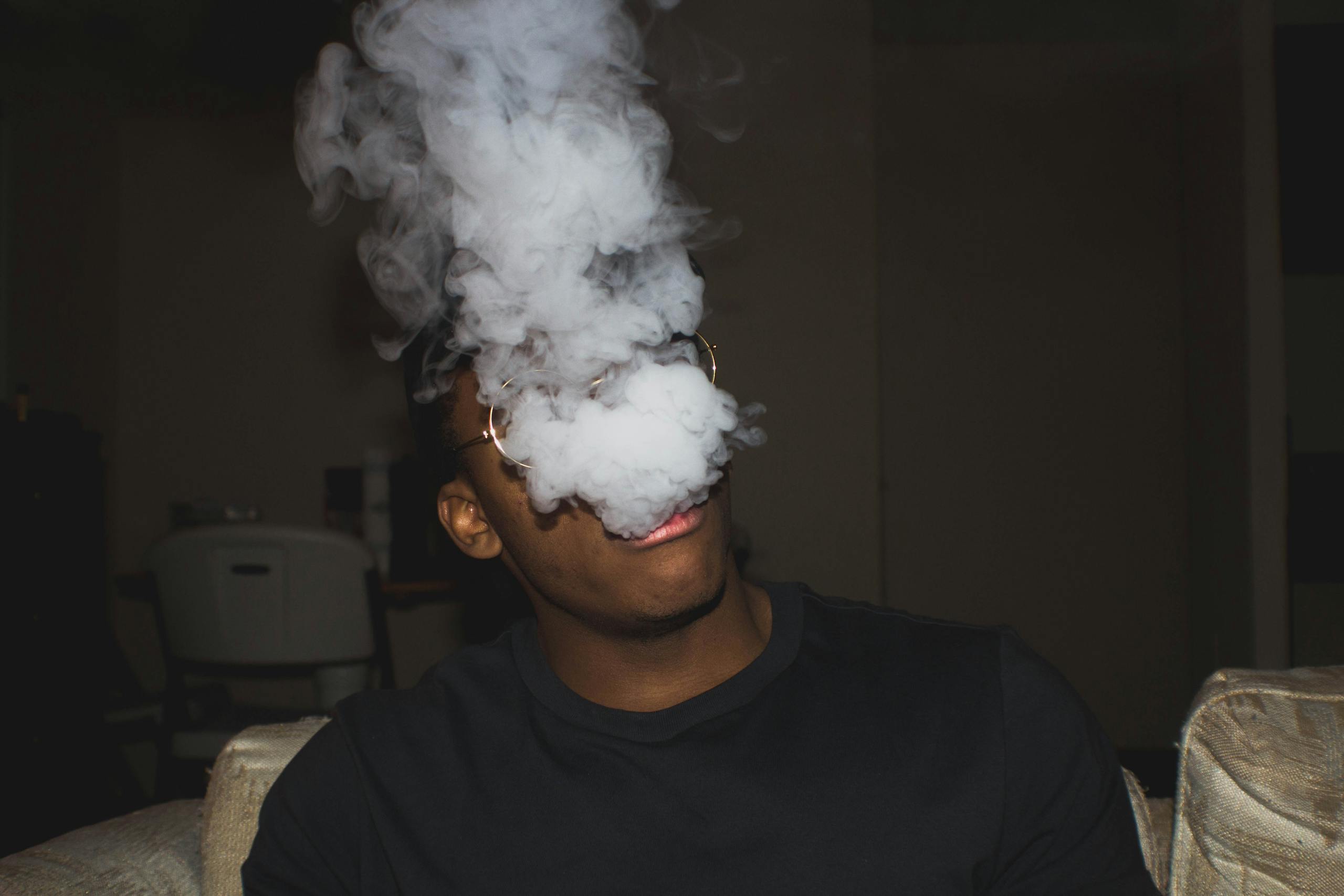 Black man sitting on sofa exhaling thick vapor, captured indoors with moody lighting.